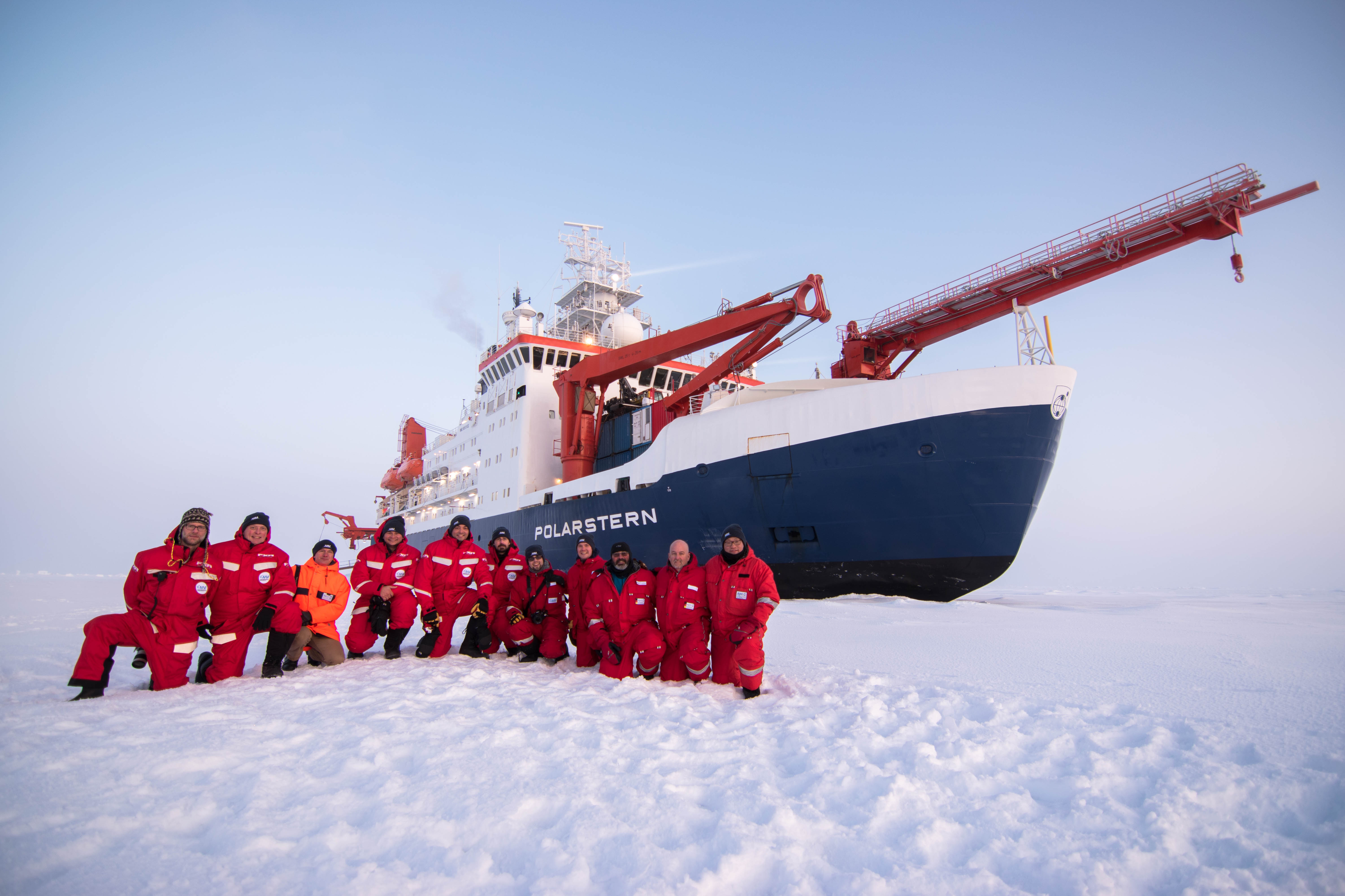 MOSAiC installation team members kneel in snow in front of the icebreaker R/V Polarstern