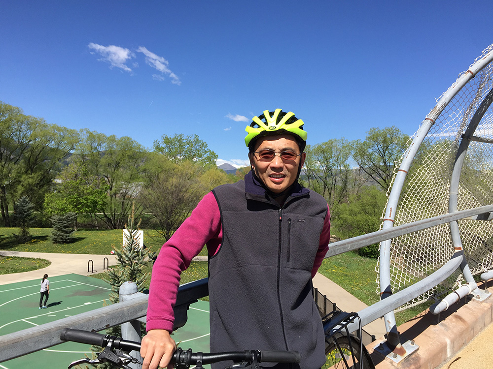 Zhien Wang stops with his bicycle to take a picture on a bridge overlooking a park and basketball court.
