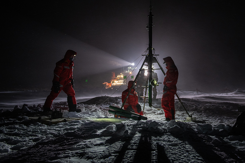MOSAiC personnel, suited up in snowsuits and headlamps, work during polar night to set up a 30-meter tower on ice and snow.