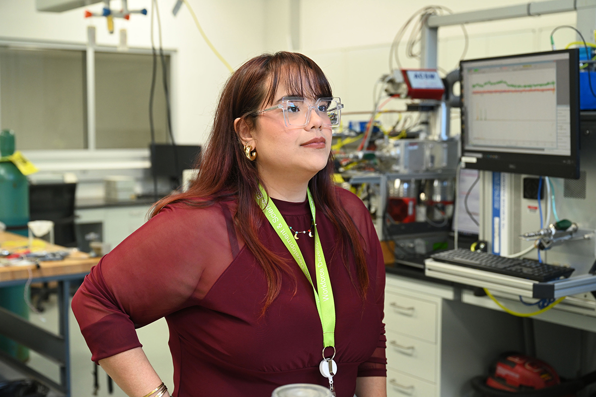 A woman in a red blouse, clear, square glasses, golden jewelry, and a green lanyard around her neck