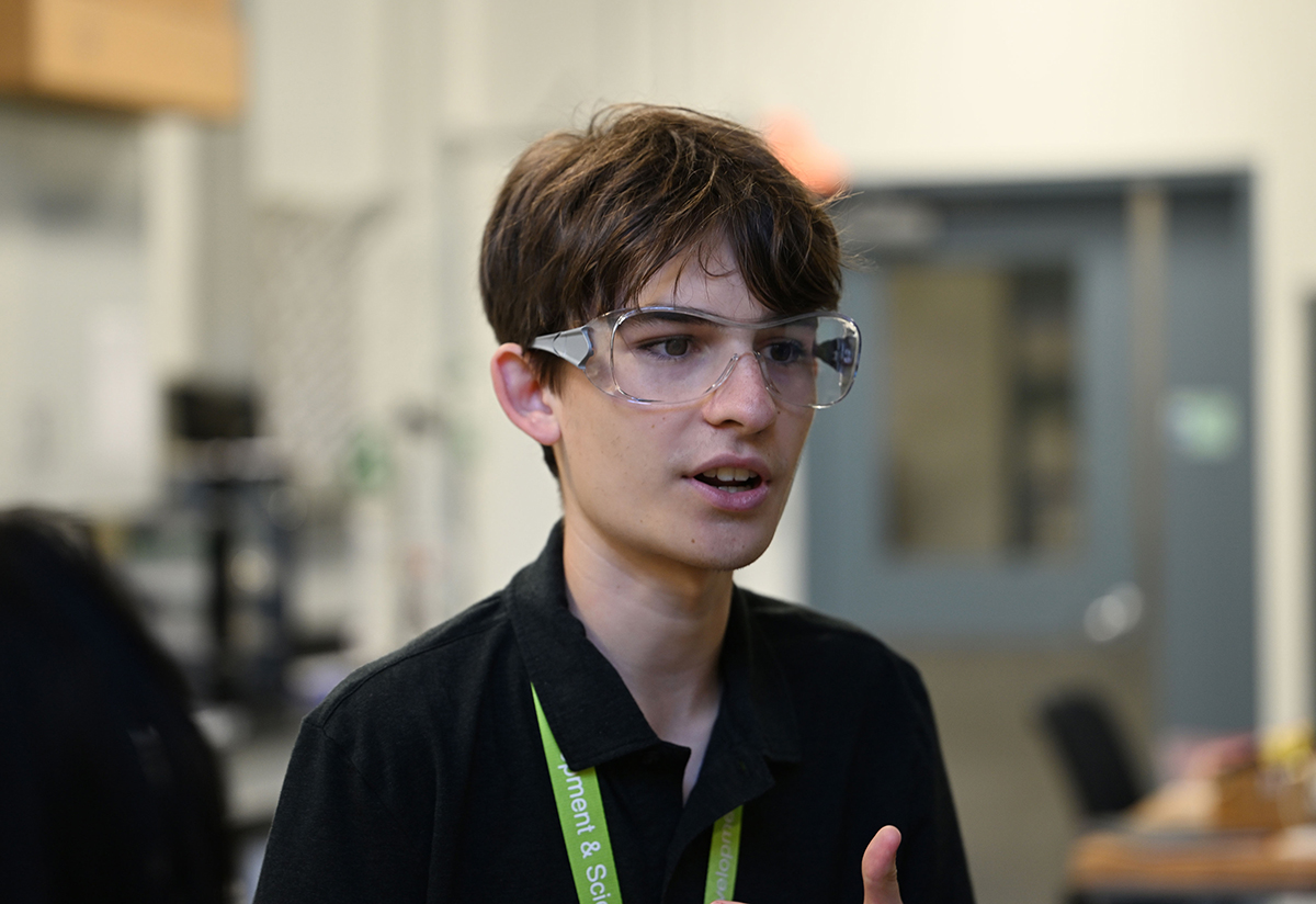 A young man in safety goggles and a collared, black shirt with a neon green lanyard around his neck