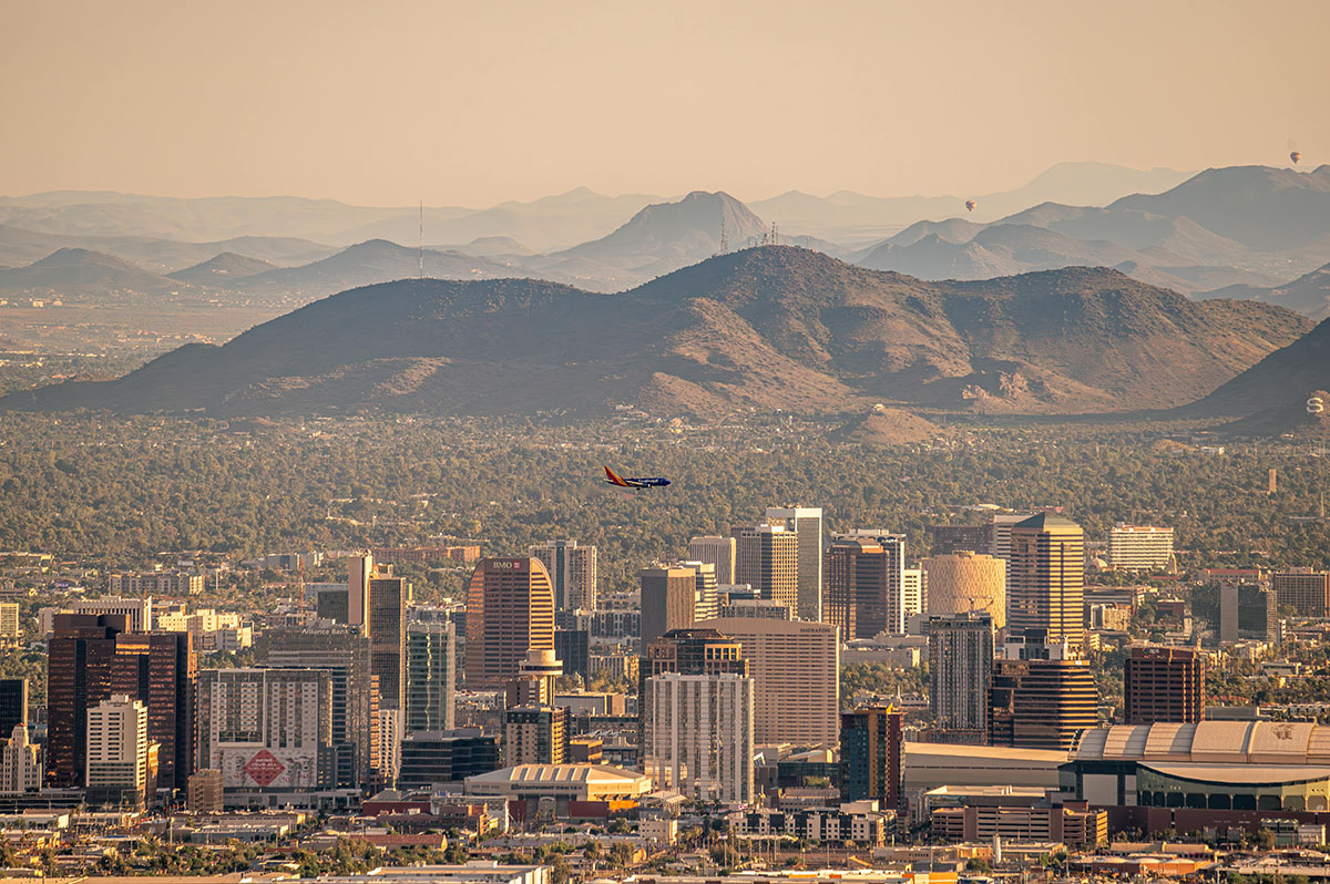 A plane flies over the downtown skyline of Phoenix, Arizona, with hot air balloons floating over distant mountains. Photo is courtesy of Arizona State University.