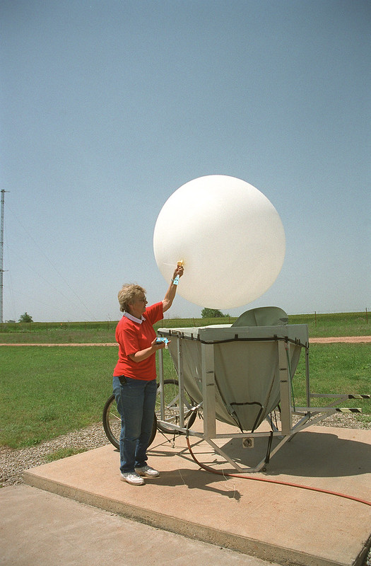 A woman wearing a red shirt and jeans holds onto the bottom of an inflated weather balloon.