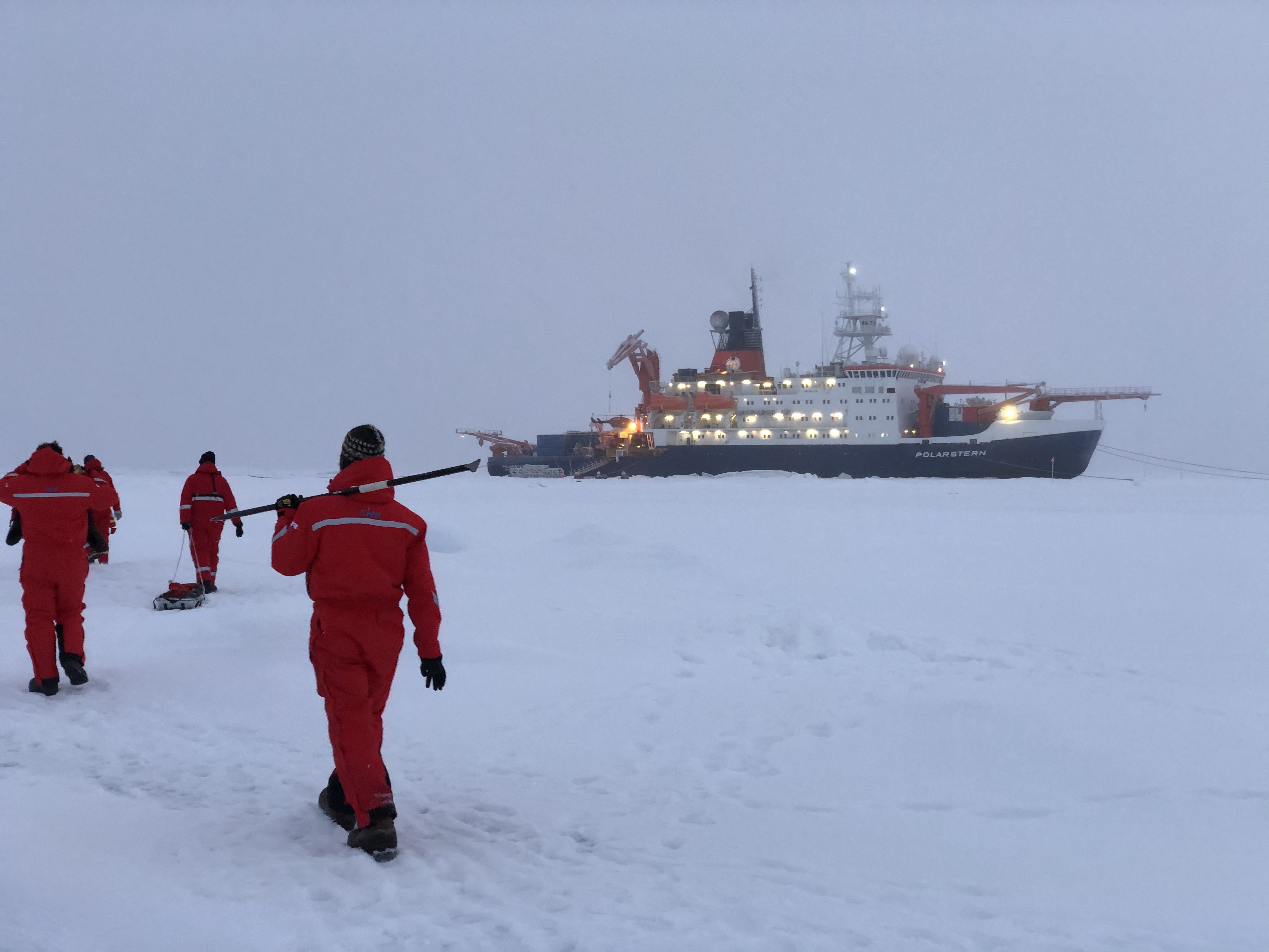 Scientists and technicians trudge through snow and ice in the central Arctic with the icebreaker Polarstern in the distance.