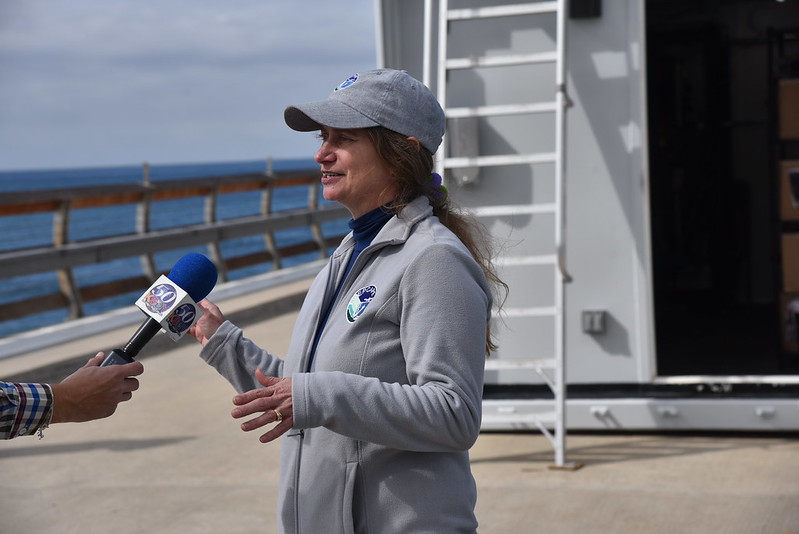EPCAPE Principal Investigator Lynn Russell speaks into a reporter's microphone in front of an ARM Mobile Facility container on the Ellen Browning Scripps Memorial Pier.