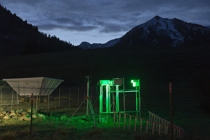 The glow of green lights from a snowflake camera illuminates the nearby radar wind profiler at the SAIL ARM Mobile Facility site in Gothic, Colorado. Snow appears on the mountain peaks in the distance with faint light peeking from behind.