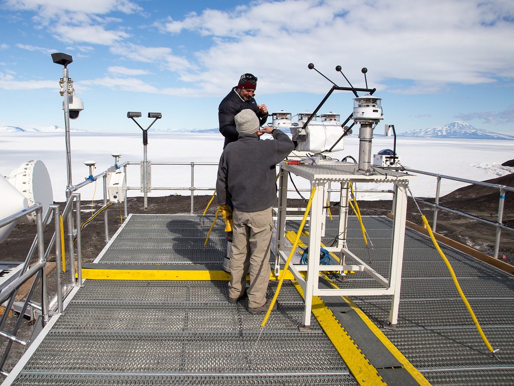 Two technicians confer next to a set of radiometers while standing on an instrument platform overlooking Antarctic ice.