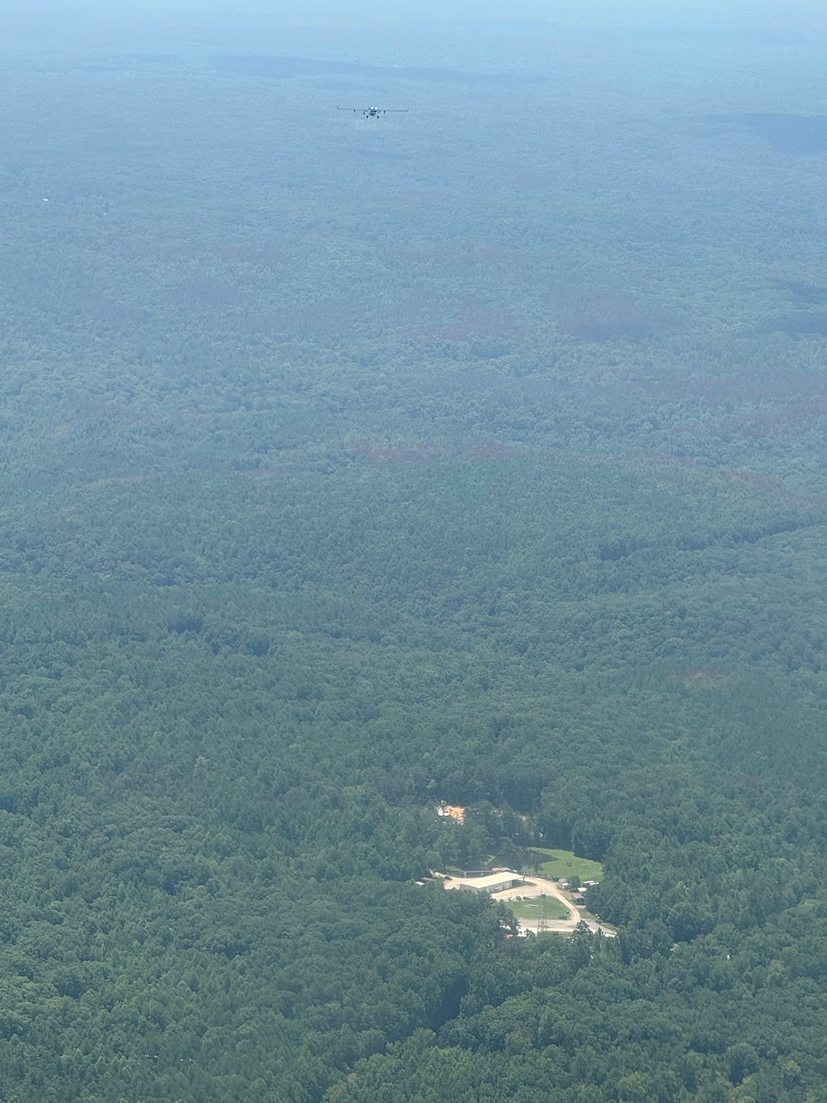 A mere speck in the air, ARM’s ArcticShark uncrewed aerial system flies over a vast forest. The only visible clearing is the one in which the Black Warrior Work Center and ARM's Bankhead National Forest atmospheric observatory are located. Photo is by Mike Hubbell, Pacific Northwest National Laboratory.