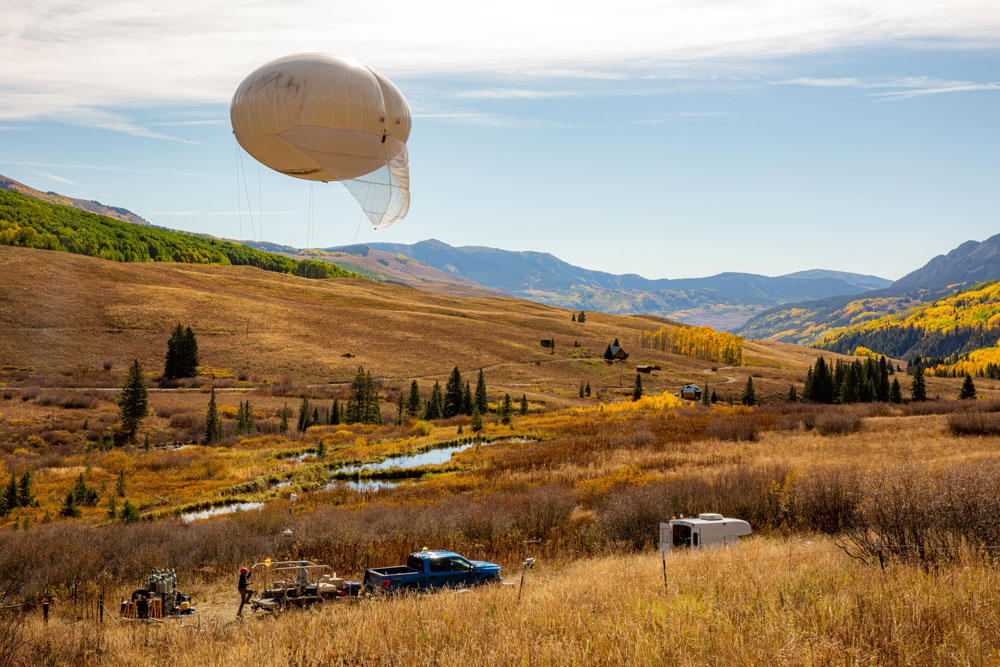 A tethered balloon is aloft over the foothills of Gothic Mountain. The landscape is mostly brown with some green along the mountainside.