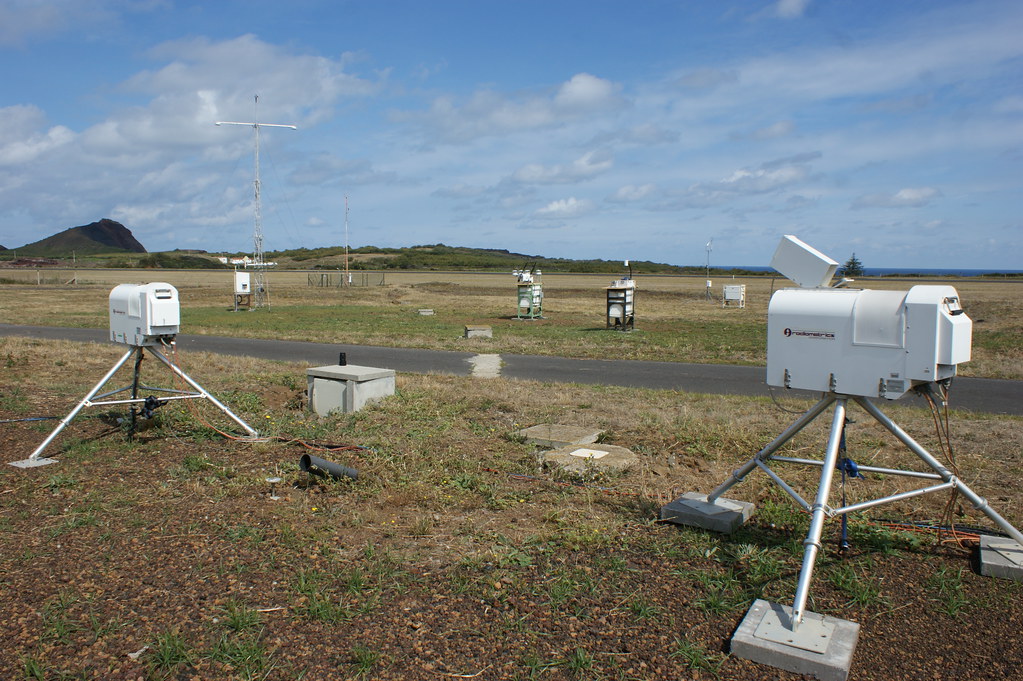 A concrete path cuts through two fields. Microwave radiometers stand on tripods in a sparsely grassy field, and more instruments operate in the field on the other side of the path.