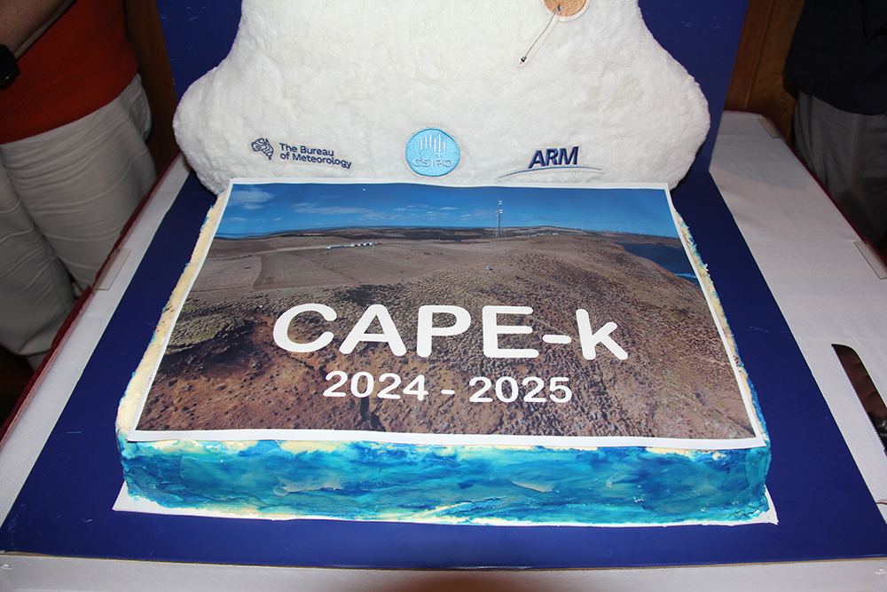An aerial photo of kennaook / Cape Grim, Tasmania, and the CAPE-k site adorns the top of a cake. The picture is labeled with the CAPE-k acronym and the campaign years of 2024 - 2025. The sides of the cake are bright blue. A weather balloon decoration and the logos of BOM, CSIRO, and ARM are attached to a puffy cloud at the top edge of the cake. Photo is by Sophie Schmidt, CSIRO.