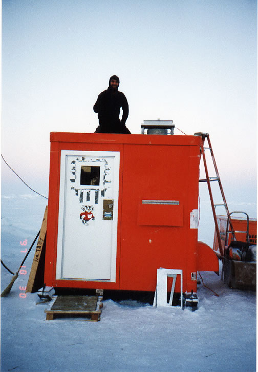 Wearing all black, Connor Flynn kneels on top of an orange instrument shelter standing on ice. The shelter door is white with a sticker of University of Wisconsin mascot Bucky Badger.