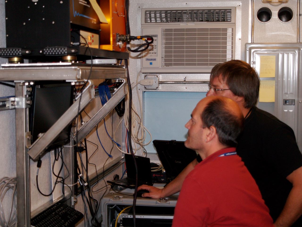 Connor Flynn sits next to Luc Rochette, who is moving a computer mouse. Both are looking at a screen mounted on the wall.