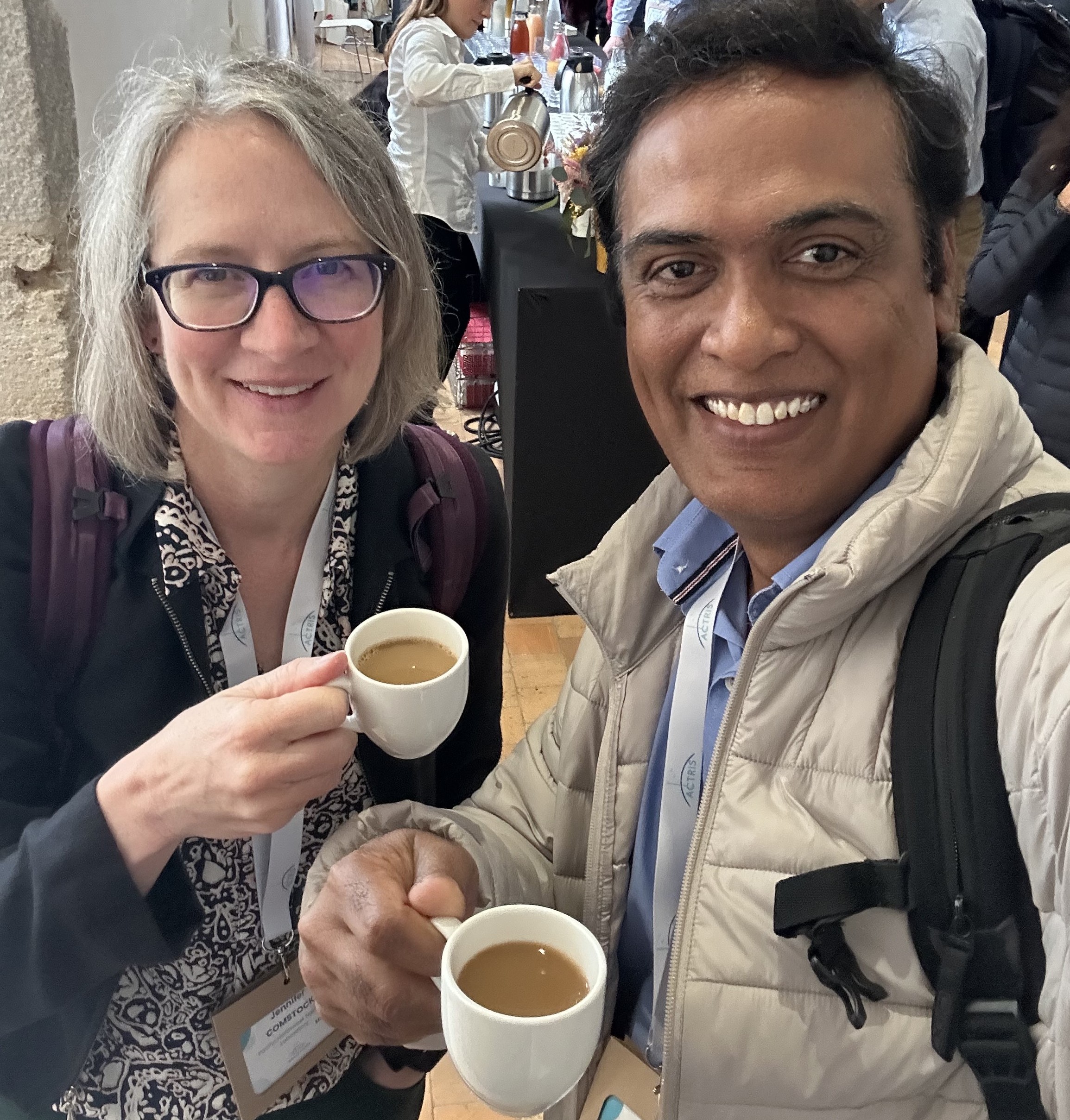 Jennifer Comstock and Giri Prakash, who are each holding a mug of coffee in their right hand, huddle together and smile in the frame.
