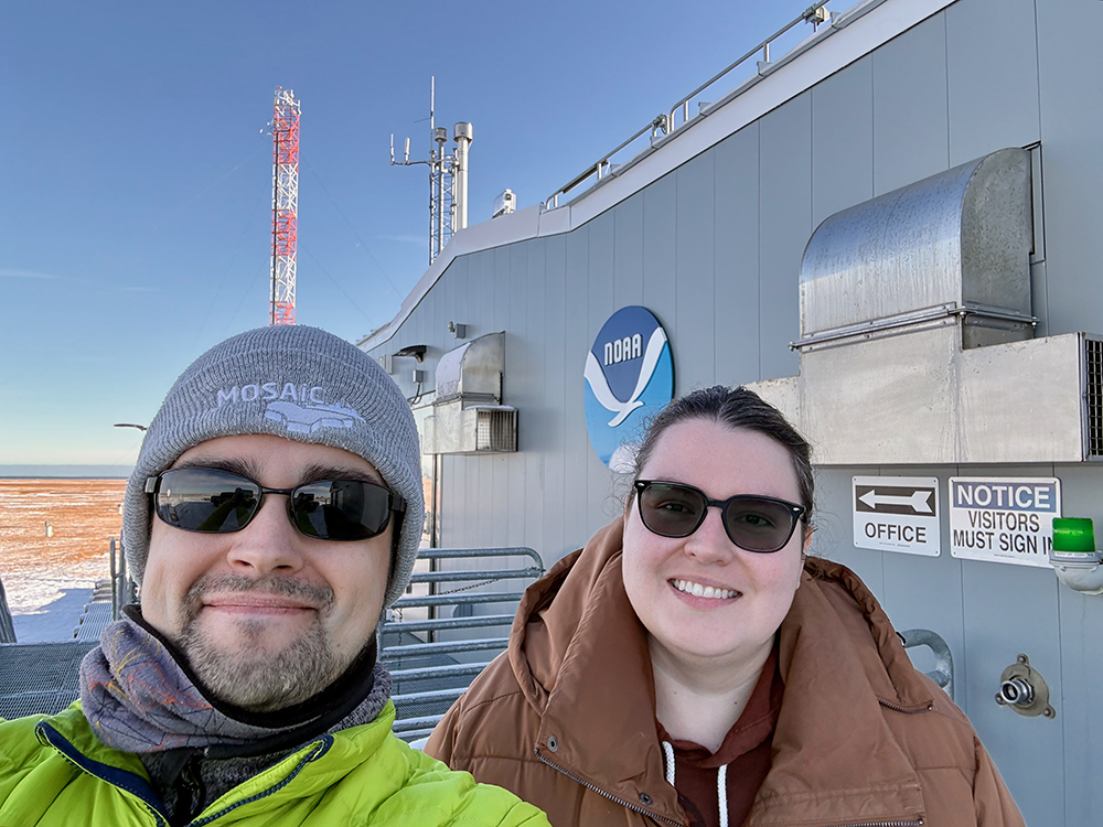 Janek Uin and Maria Zawadowicz smile at the camera while standing outside a building with the NOAA logo on the front.