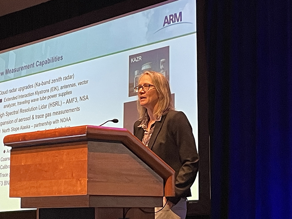 A picture of a Ka-Band Zenith Radar (KAZR) is visible on a screen behind Jennifer Comstock as she presents at a podium.