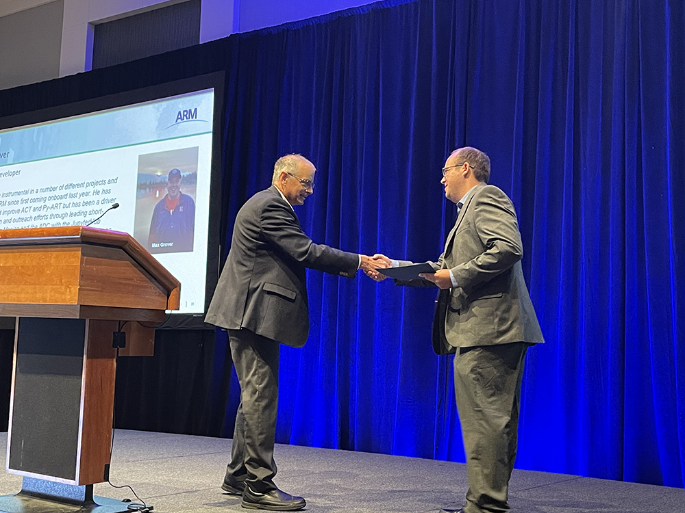 Jim Mather and Max Grover shakes hands onstage. Behind Mather is a large monitor showing a presentation slide on Grover.