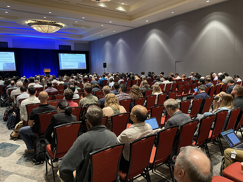 People sit in rows in a conference room with two screens mounted at the front showing a presentation on tethered balloon activities.