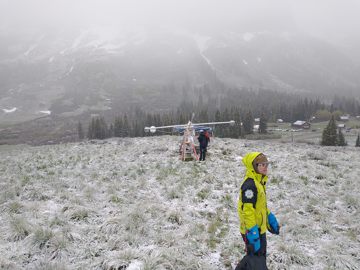 A technician wearing a bright yellow jacket, robin's egg blue gloves, and navy snowpants walks across frost- and snow-covered grass with colleagues working on instrumentation over to his left.