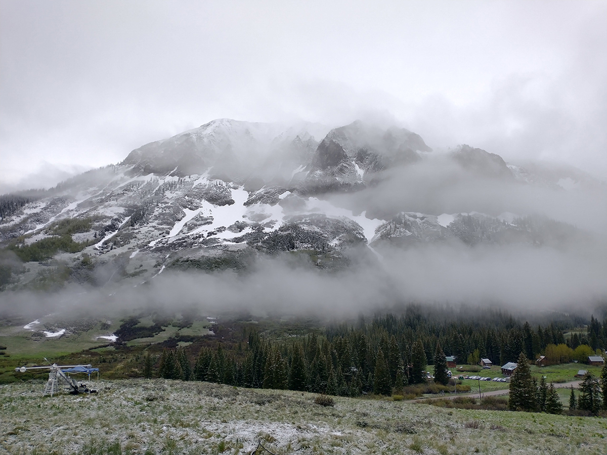 Low clouds settle over a patch of trees at the base of a snow-covered mountain.