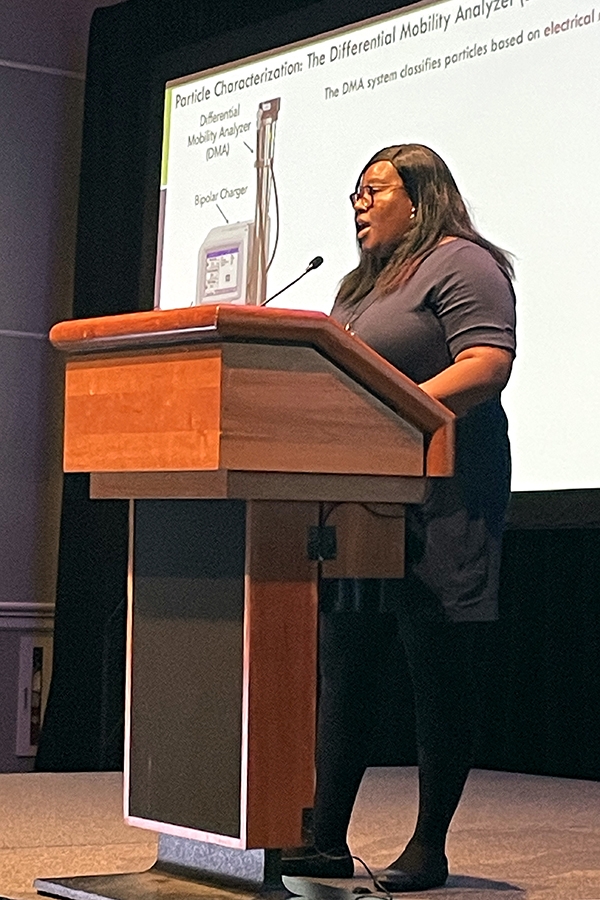 Ogo Enekwizu speaks into a microphone at the podium with a slide behind her showing a differential mobility analyzer and bipolar charger. The slide says that the DMA system classifies particles based on electrical mobility.