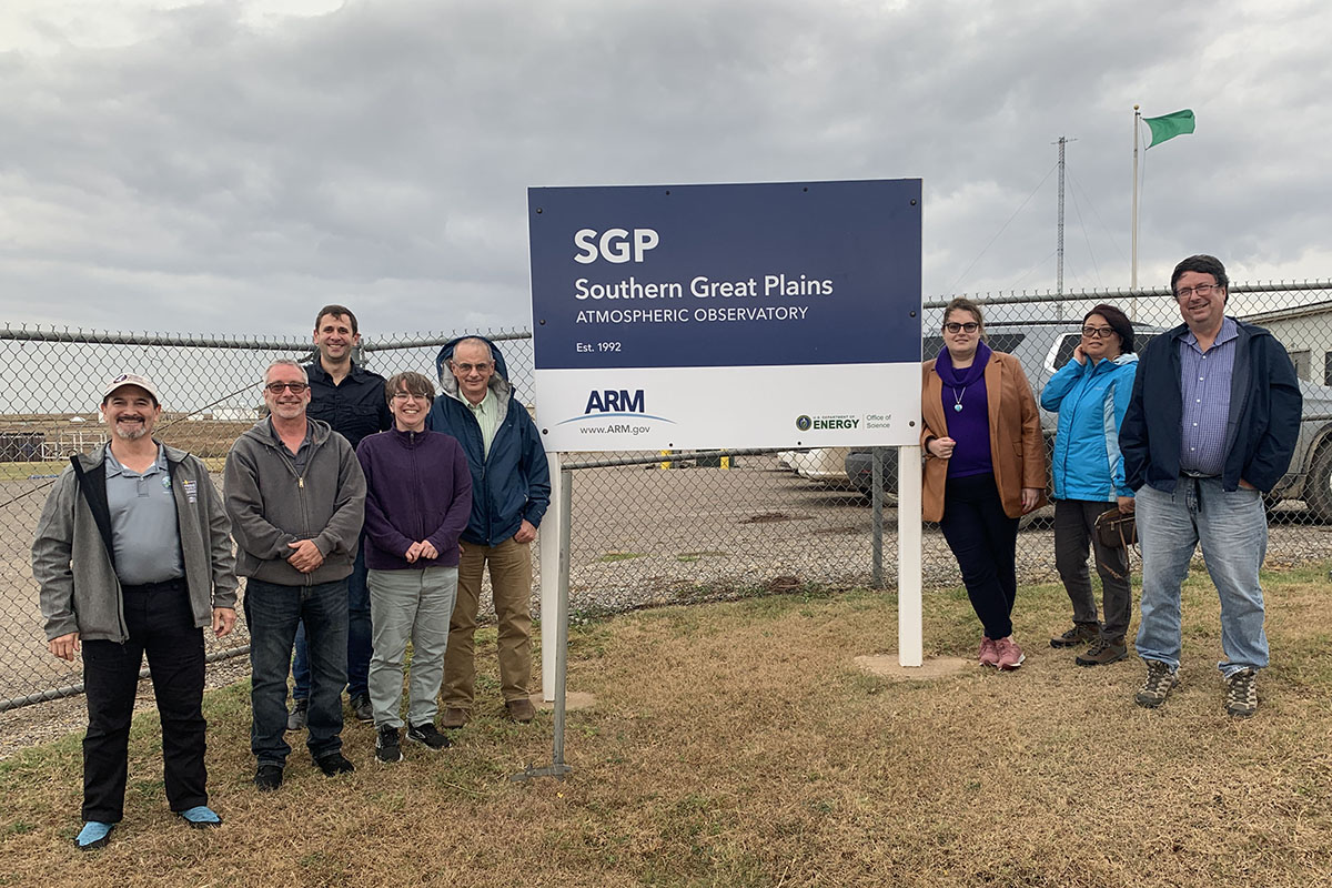UEC workshop attendees flank the sign greeting visitors at the entrance of the Southern Great Plains atmospheric observatory. Five people are on the left, and three are on the right.
