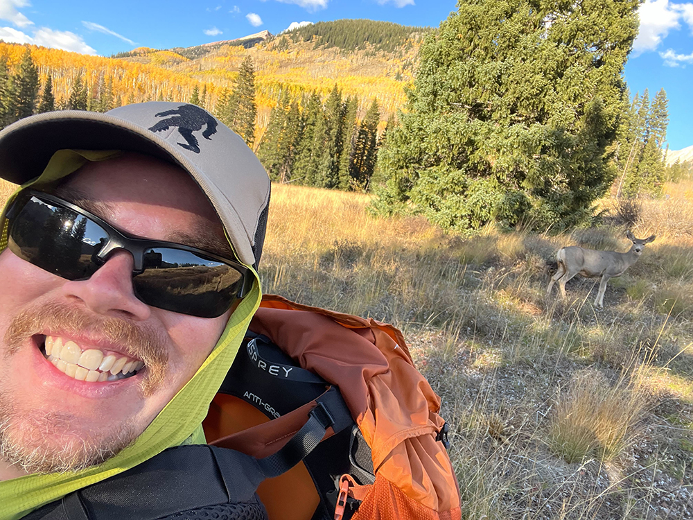 Aaron Kennedy looks to his left and smiles for a picture as a deer stops behind him and looks at the camera.