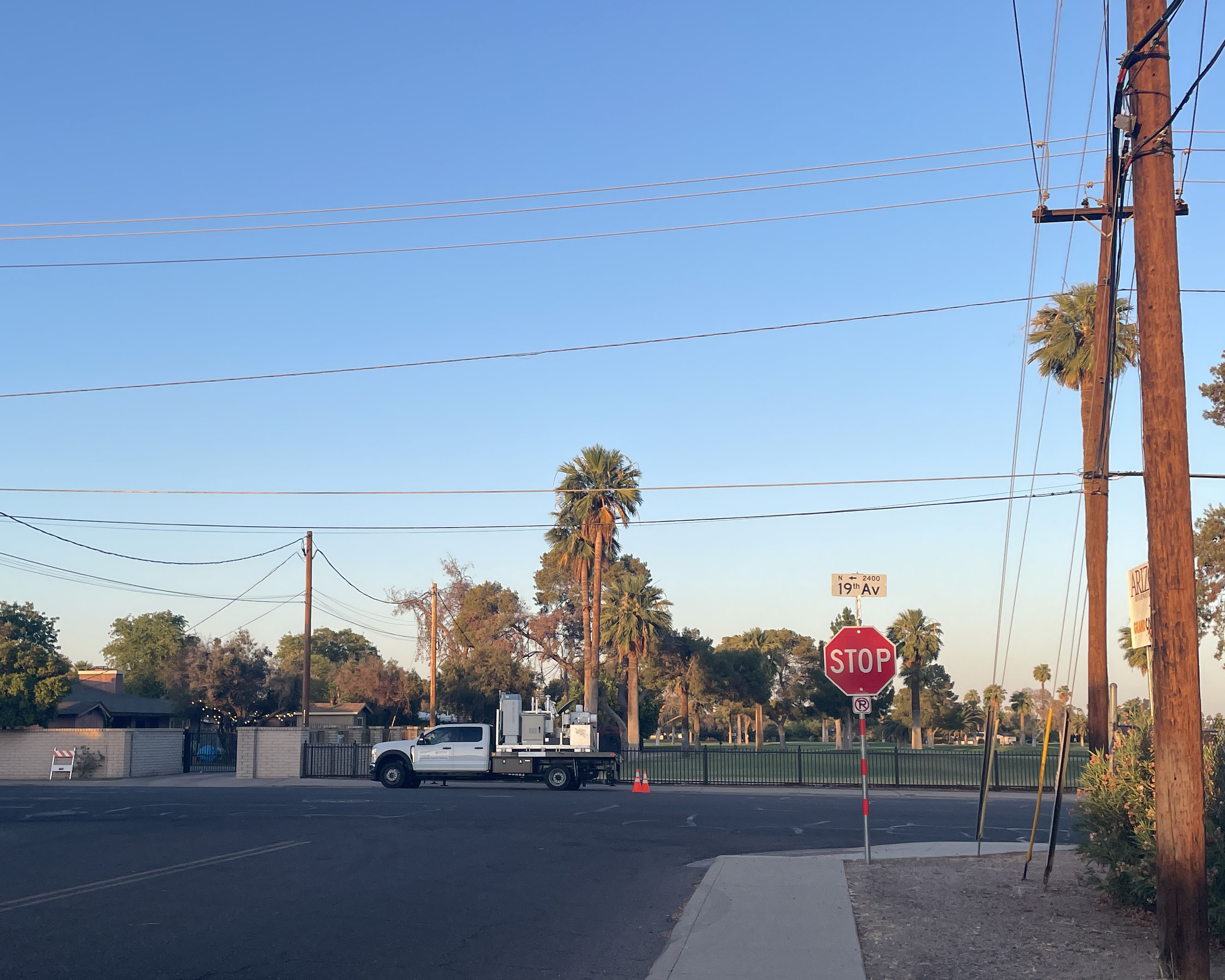 A truck with instruments is parked along a street next to a fence with grass and trees beyond it. A stop sign and street sign with 19th Av are in the foreground.