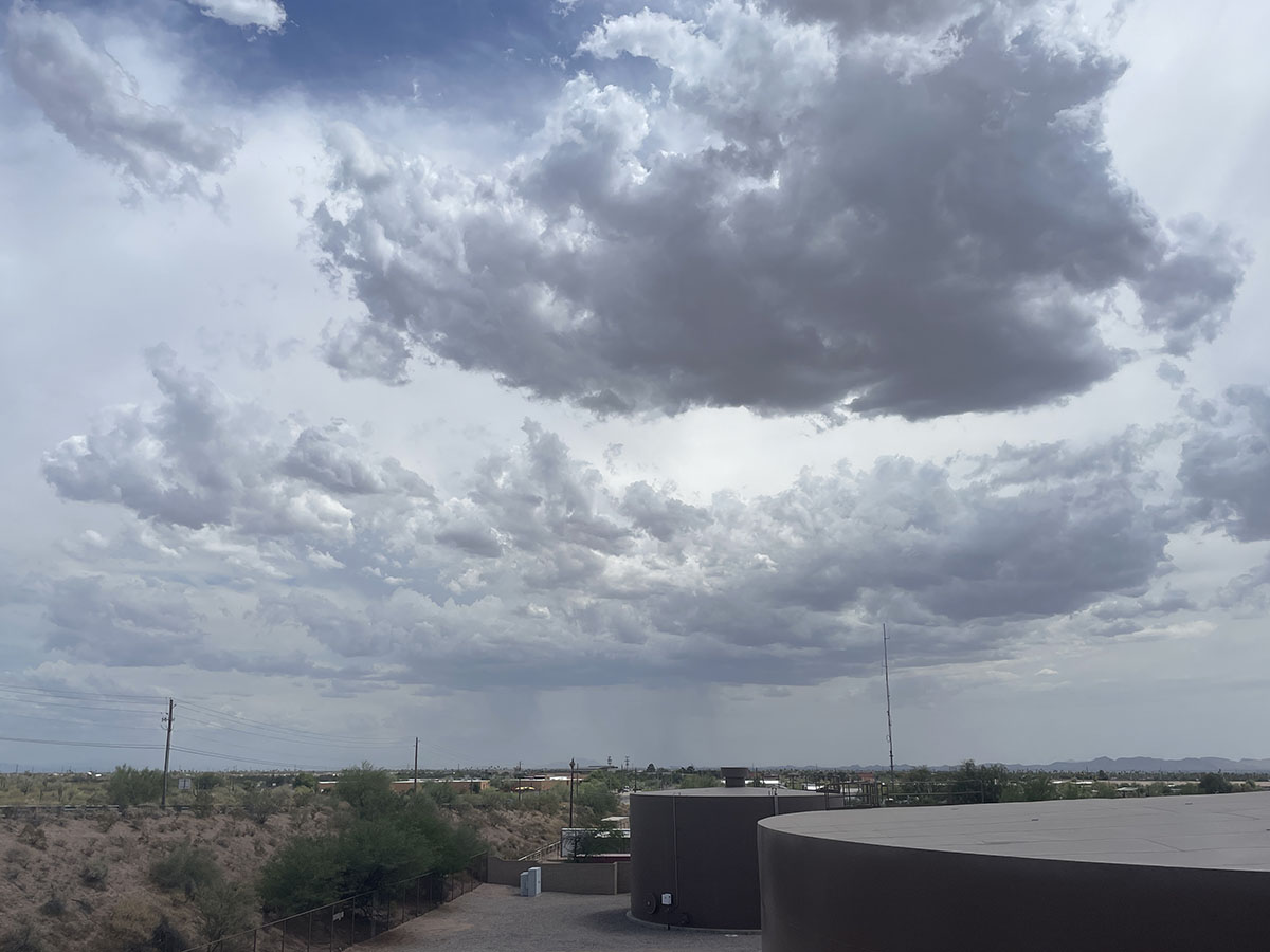 Rain clouds float above a desert landscape with some vegetation and buildings. Streaks of rain are seen in the distance.