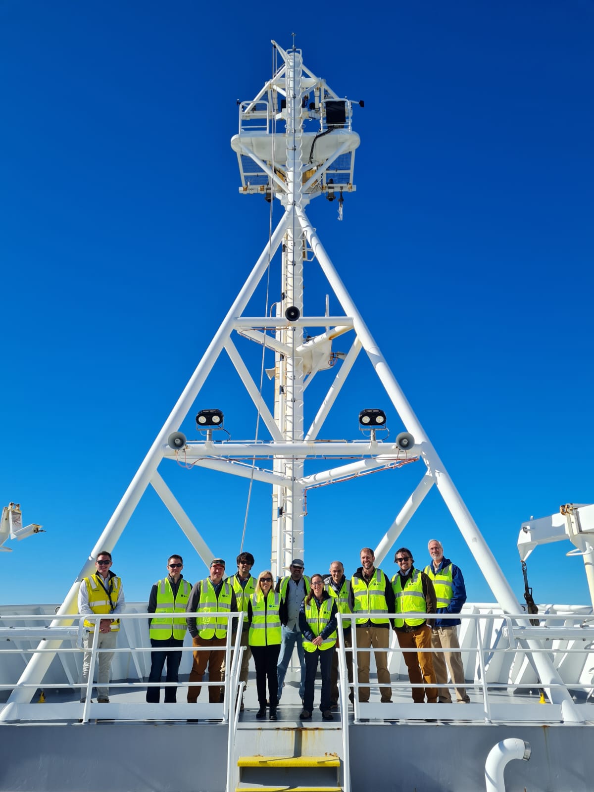 A brilliant blue sky over Hobart, Tasmania, serves as a vibrant backdrop for a tour of the CSIRO-operated research vessel Investigator, which will collect data off the coast of the CAPE-k site in May 2025. A group of visitors on the ship pose for a photo. Photo is by Sally Taylor, CSIRO.
