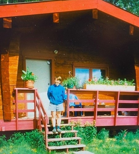 Jen Delamere stands on the stairs of her cabin, with plants lining the deck railing next to her.