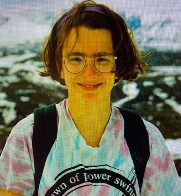 Jen Delamere smiles for the camera while wearing a tie-dyed T-shirt, glasses, and a backpack.