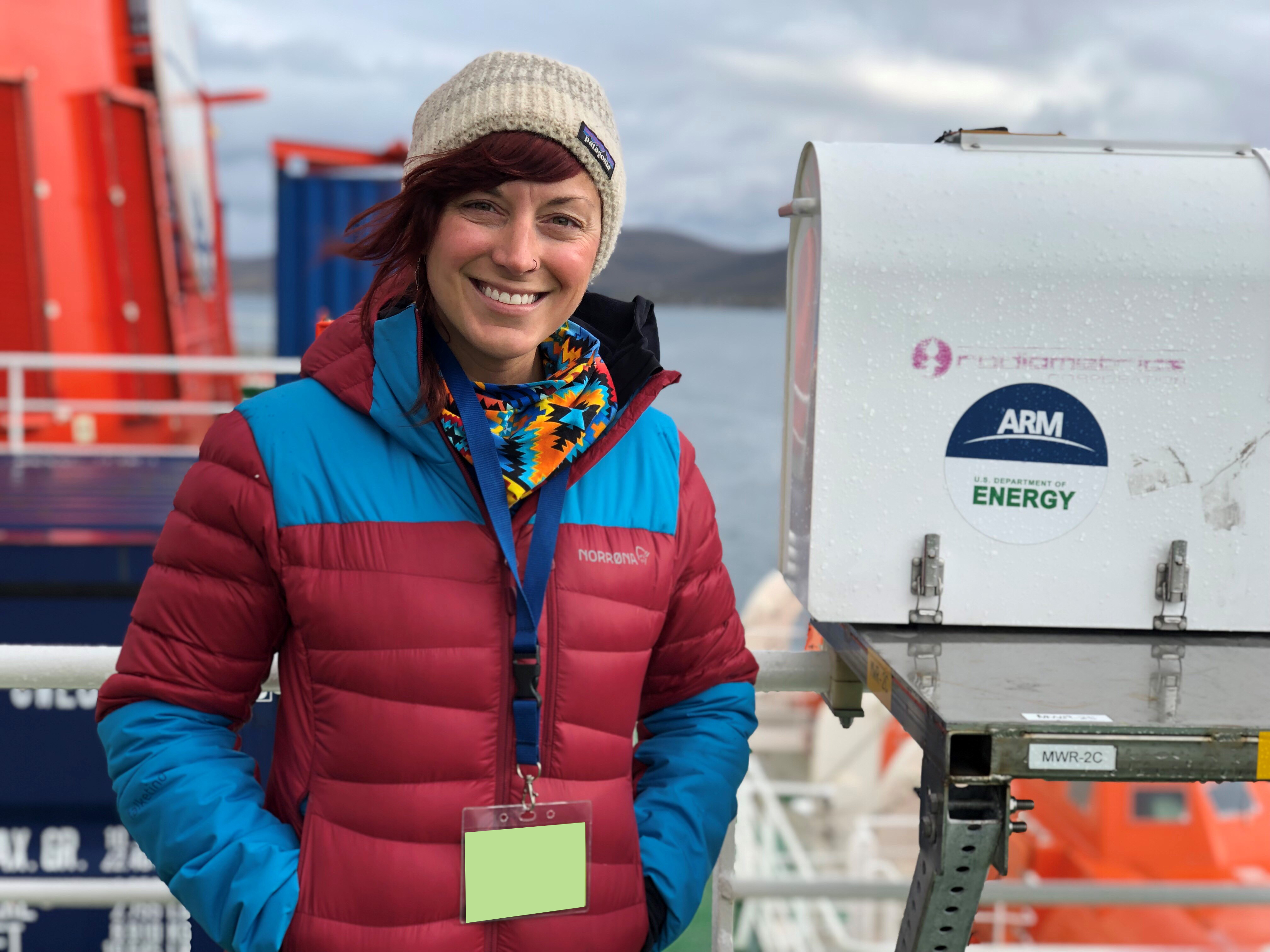 Wearing a red-and-blue jacket and beige beanie, Jessie Creamean stands next to a 3-channel microwave radiometer before embarking on the MOSAiC expedition in 2019.