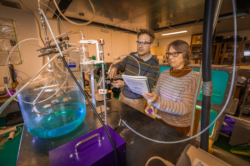 Wearing safety glasses and surrounded by tubing, Daniel Knopf and Josephine Aller work together in a lab.