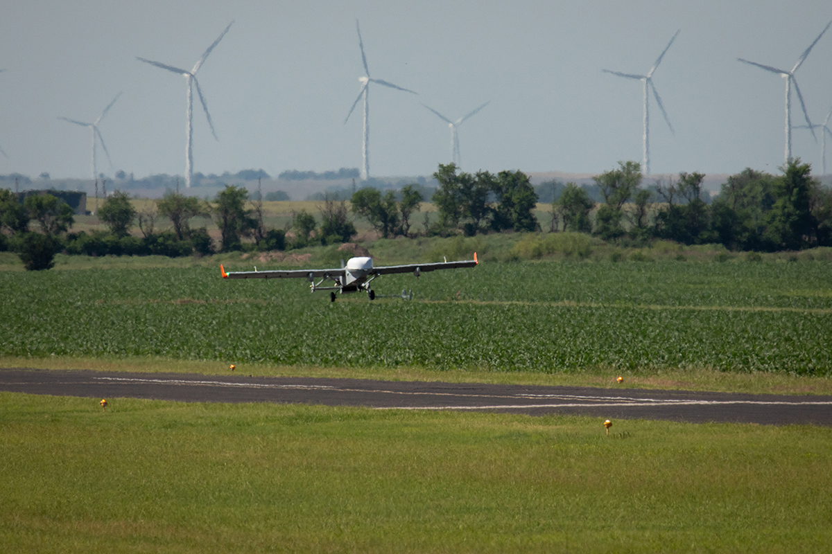 With wind turbines in the distance, the ArcticShark uncrewed aerial system is captured over the runway at the Blackwell-Tonkawa Municipal Airport.