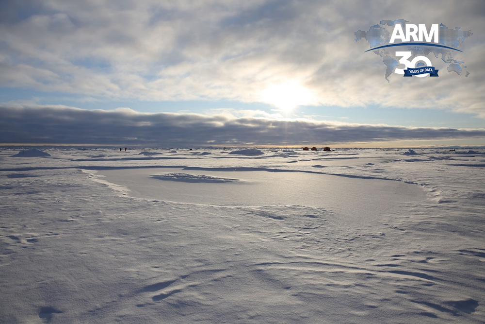 A streak of blue sky breaks up the clouds hovering over the central Arctic ice and snow. Researchers and shelters are seen in the distance.