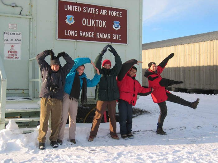 Five researchers wearing winter jackets, pants, and boots use their bodies to spell out "M-PACE" in front of a building with a sign that says, "United States Air Force Oliktok Long Range Radar Site."