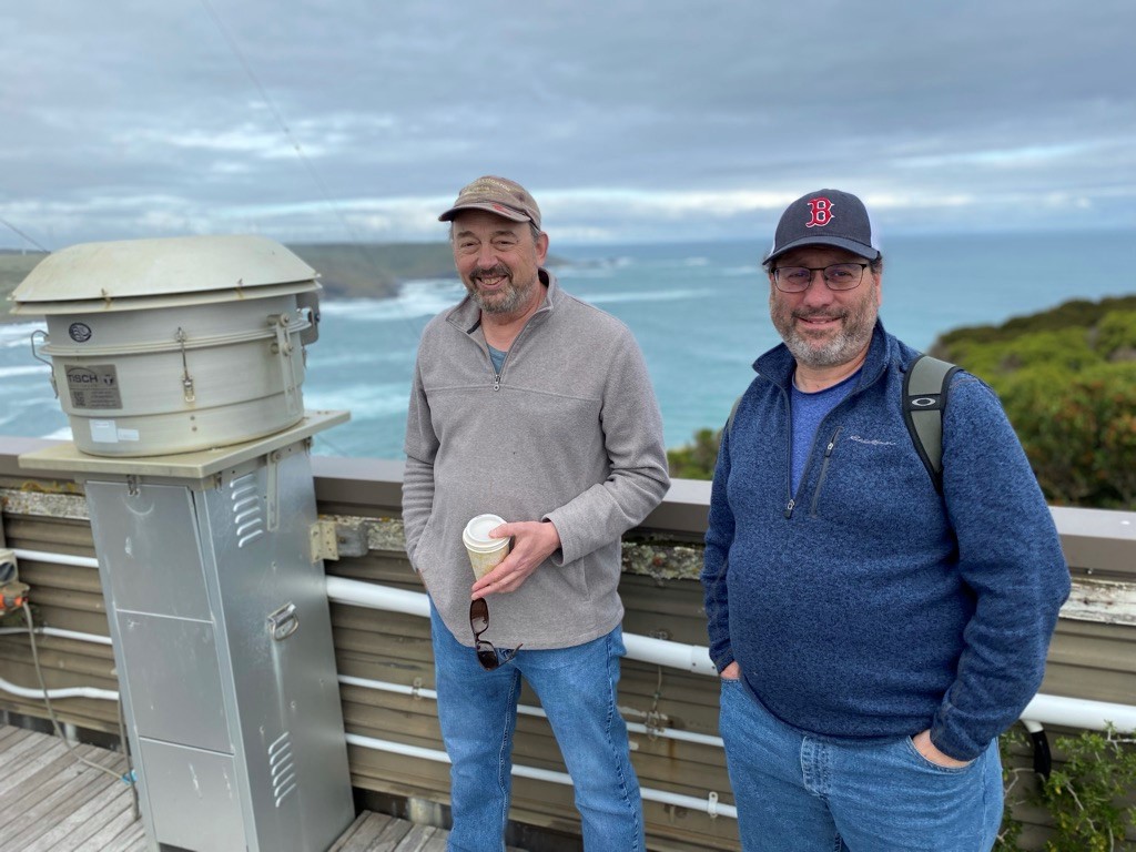 Jay Mace holds a coffee cup in his hand while standing next to Roger Marchand, who is also wearing a ballcap on a cloudy day in northwestern Tasmania.