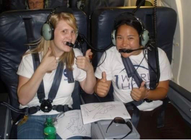 Christina McCluskey and Jennifer DeHart give thumbs up for the camera. They are wearing headsets and are strapped into their seats aboard the aircraft.