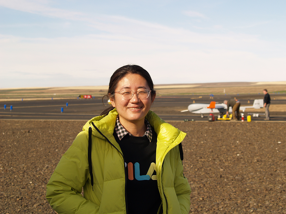 Fan Mei smiles for the camera while crew members work on the ArcticShark uncrewed aerial system behind her.