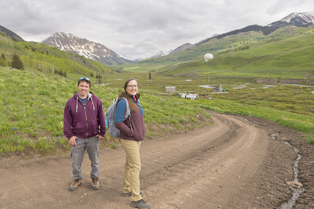 Daniel Feldman and Allison Aiken stand on a dirt road and smile at the camera, with the tethered balloon system behind them.