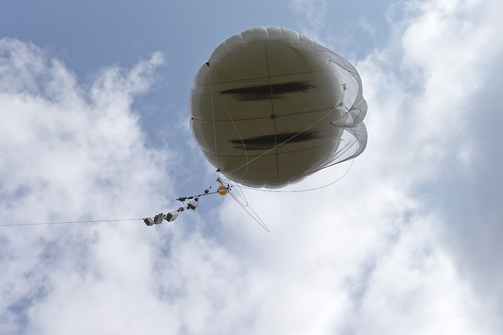 With clouds above, a tethered balloon is aloft with a string of instruments hanging below.
