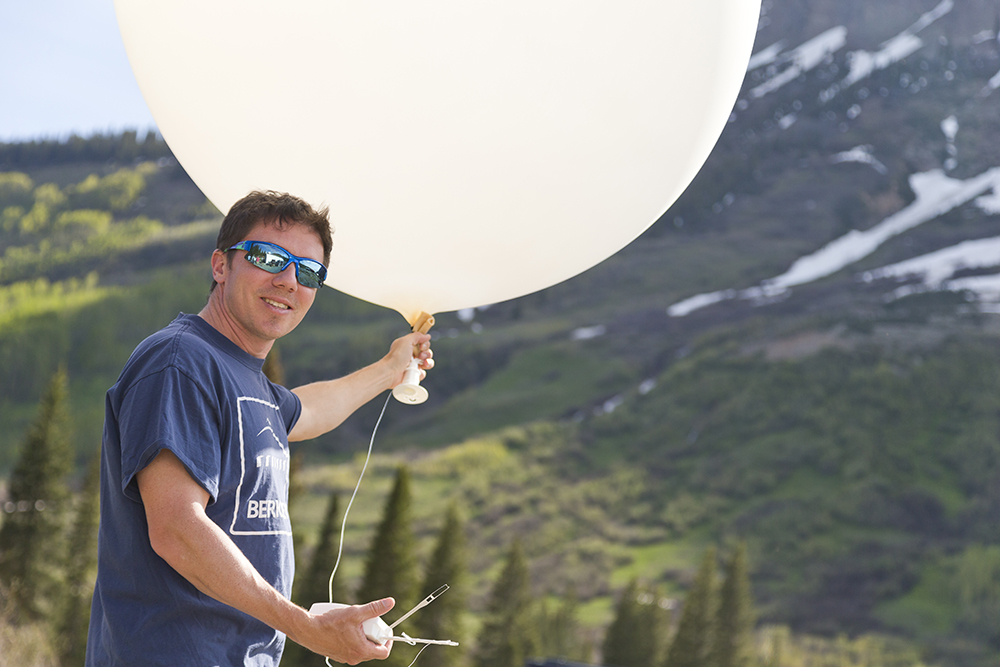 Wearing sunglasses and a Berkeley Lab T-shirt, Daniel Feldman looks at the camera while holding the bottom of a weather balloon in his left hand and a radiosonde in his right hand. Trees and snow on a mountainside are visible in the distance.