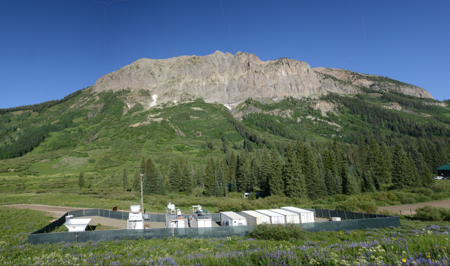 ARM Mobile Facility containers and instruments are situated inside a fence. The photo, taken on a sunny day, also shows a mountain with patchy snow and trees populating the lower half of the mountainside.