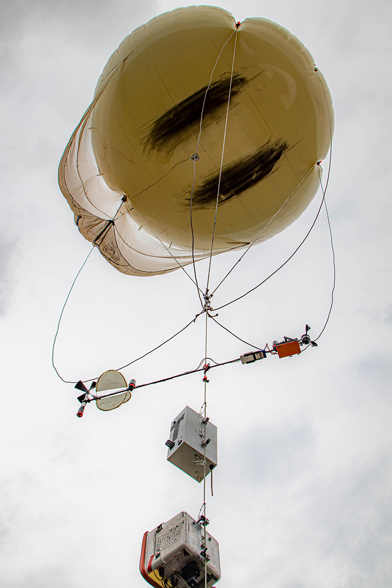 A tethered balloon system, with instruments strung along the tether, is aloft in a cloudy sky. 