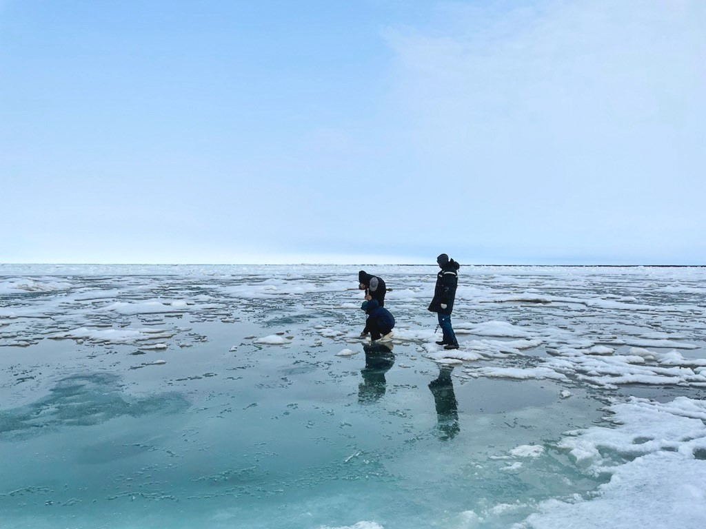 Three people wearing jackets, snow pants, and boots stand on a patch of slushy sea ice.