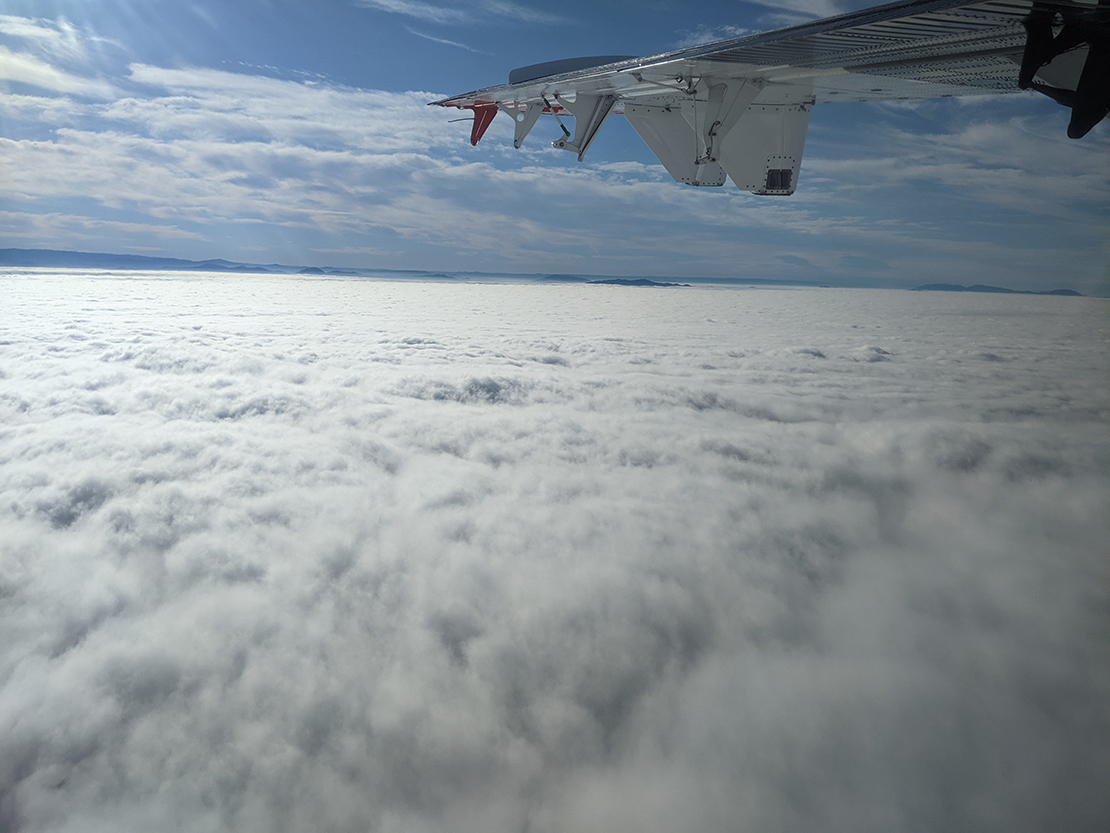 A wing of the Twin Otter hovers above a cloud bank. Mountain peaks rise in the distance. Higher-level clouds are seen above the plane.