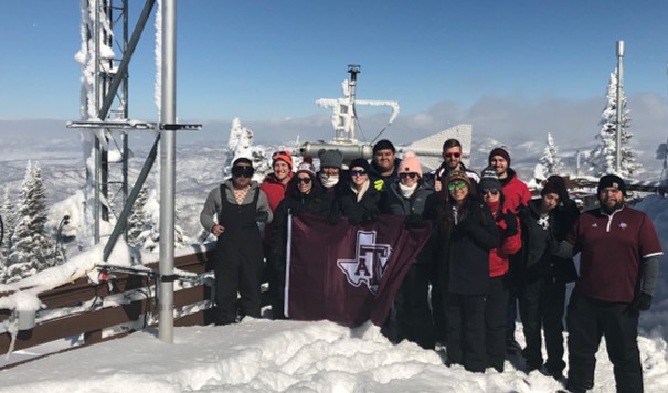 Sarah Brooks stands with students at snow-covered Storm Peak Laboratory, with those in front holding a Texas A&M flag.