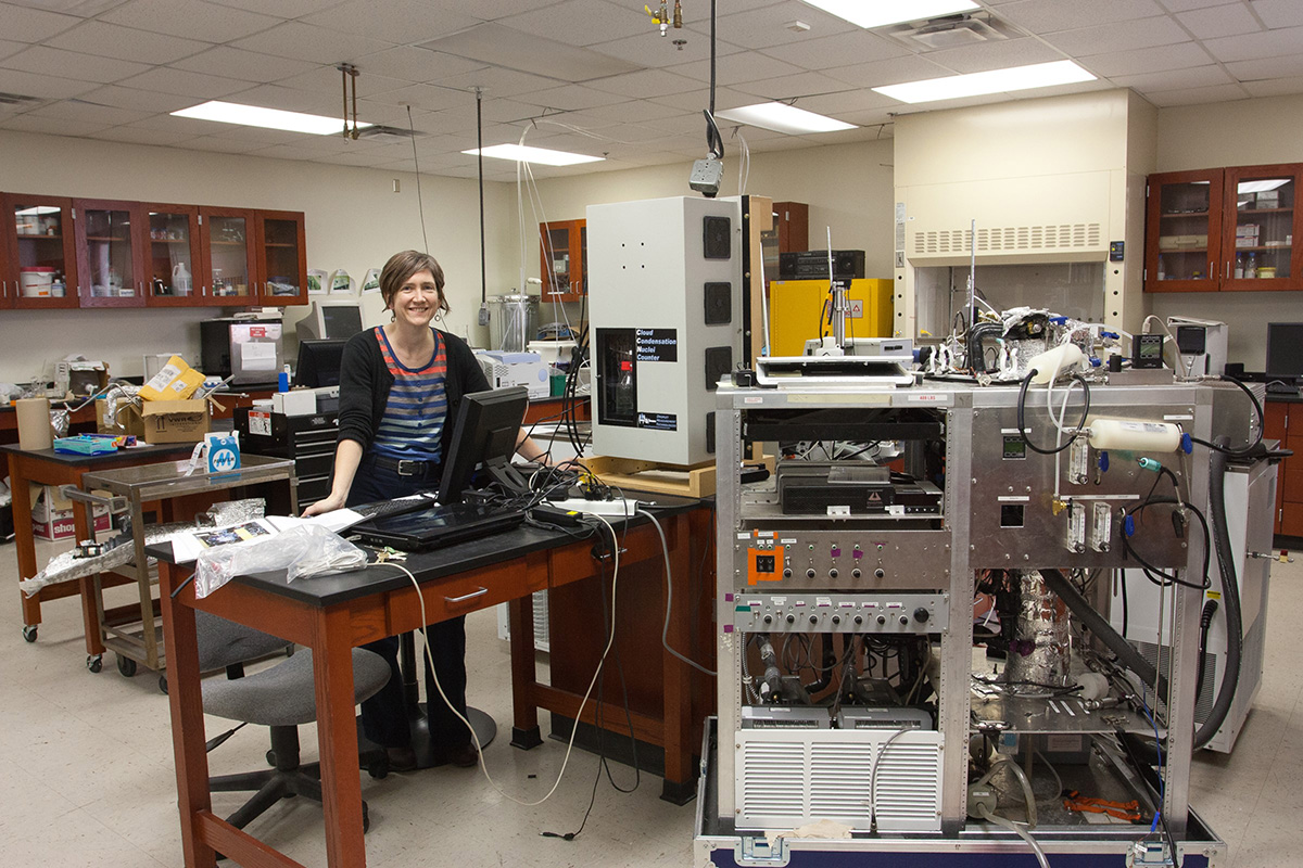 Sarah Brooks stands behind a table with a computer on top, a cloud condensation nuclei counter to her left, and an instrument system on a large metal cart in front of her.
