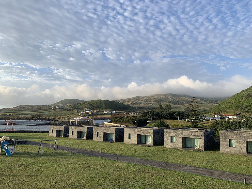 Stratocumulus clouds stretch over the hills of Graciosa Island. A playground stands in front of a row of six gray hotel villas, each with one small window and one larger window opening to the outside.